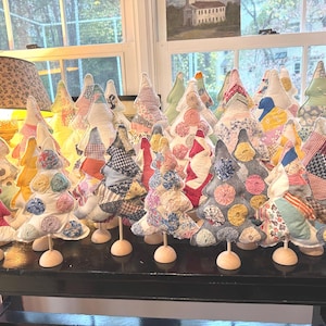 Könnte beinhalten: Eine Sammlung handgefertigter Stoff-Weihnachtsbäume in verschiedenen Größen und Mustern. Die Bäume bestehen aus Patchwork-Stoffen in Rot, Grün, Blau und Gelb und sind auf kleinen Holzsockeln montiert. Die Bäume sind auf einem schwarzen Tisch angeordnet.