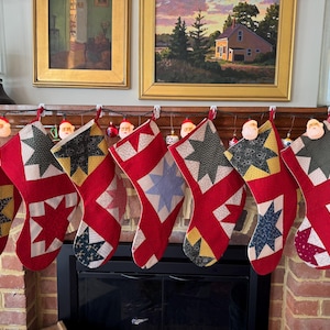 May include: A row of eight red Christmas stockings with various star patterns in yellow, green, and blue. Each stocking is decorated with a different patchwork design and hung above a fireplace. Santa Claus lights are strung above the stockings.