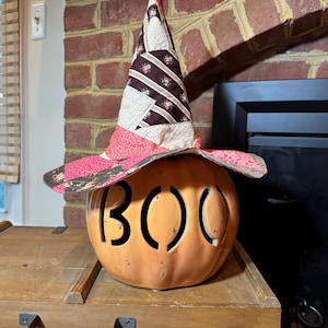 May include: A carved orange pumpkin with the word "BOO" cut out, topped with a patchwork witch hat. The hat features a mix of floral and geometric patterns in shades of pink, brown, and white. A festive Halloween decoration.