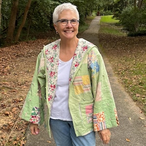 May include: A light green quilted jacket with a floral print hood and patchwork details. The jacket is worn over a white top and blue jeans. The jacket has a relaxed fit and a vintage aesthetic.
