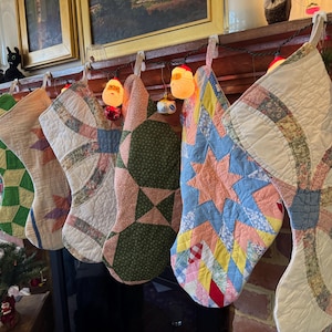 May include: A selection of handmade Christmas stockings with patchwork quilt patterns in diverse colours. The stockings are suspended from a mantelpiece adorned with lights and decorations, forming a festive holiday arrangement.