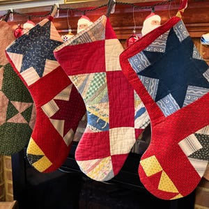 May include: Five handmade Christmas stockings with patchwork designs. The stockings feature various patterns, including stars and geometric shapes, in red, blue, green, and yellow. They are hanging from a wooden mantelpiece, decorated with Christmas ornaments.