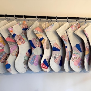 Pode incluir: Dez meias de Natal brancas com desenhos florais de patchwork. Penduradas numa barra preta, cada meia apresenta um padrão floral único com tecidos rosa, azul e amarelo. Uma decoração de Natal artesanal.