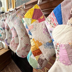 Puede incluir: Una colección de calcetines navideños hechos a mano, cada uno elaborado con tela de edredón de retazos. Los calcetines presentan una variedad de colores y patrones, incluyendo estampados florales, formas geométricas y telas lisas. Cuelgan de una repisa de madera.