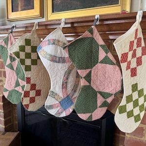 Puede incluir: Cinco calcetines navideños acolchados con varios patrones, incluyendo formas geométricas verdes y rosas, y diseños de tablero de ajedrez rojos y verdes. Los calcetines cuelgan de una repisa de madera sobre una chimenea de ladrillo.