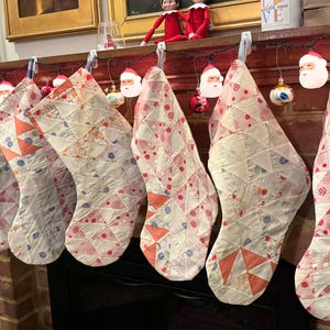May include: A row of Christmas stockings with a patchwork design in red, white, and blue hang above a fireplace. The stockings are decorated with geometric patterns and small red dots. Santa Claus ornaments and Christmas lights are also visible.