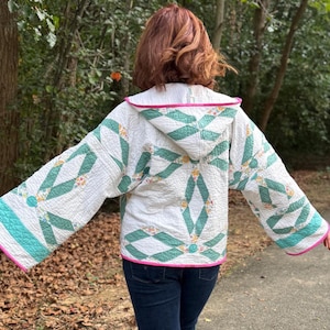 Puede incluir: Chaqueta acolchada blanca con capucha, con un patrón de diamantes verdes y blancos. La chaqueta tiene detalles turquesa en las mangas y un ribete rosa. La persona lleva vaqueros azules.