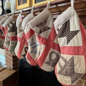 Puede incluir: Siete calcetines navideños con diseño de patchwork. Cada calcetín tiene un fondo rojo con patrones beige, marrón y azul. Cuelgan de una repisa de madera, creando una decoración festiva para la temporada navideña.