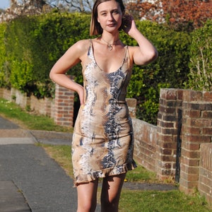 May include: A woman wearing a short, brown and black snakeskin print dress with a ruffled hem. She is wearing black ankle boots and a silver necklace.