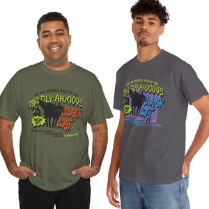 K&ouml;nnte beinhalten: Zwei Personen tragen T-Shirts mit einem grafischen Design. Das Design zeigt den Text "AND THE MYTHICAL BELLY OF THE MILDLY ANXIOUS BLACK CAT" mit einer schwarzen Katzenillustration. Die Shirts sind olivgr&uuml;n und anthrazitgrau.