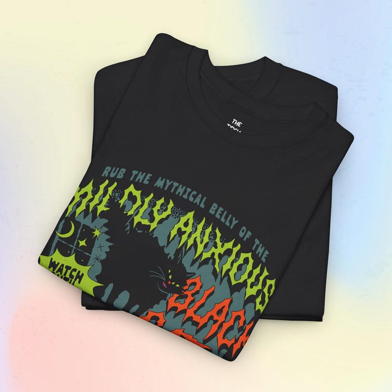 K&ouml;nnte beinhalten: Zwei schwarze T-Shirts sind &uuml;bereinander gestapelt, wobei das obere Shirt ein grafisches Design aufweist. Das Design umfasst eine schwarze Katze, einen Mond und Sterne sowie den Text "RUB THE MYTHICAL BELLY OF THE DISAVOWED". Der Text "WATCH" ist ebenfalls sichtbar.