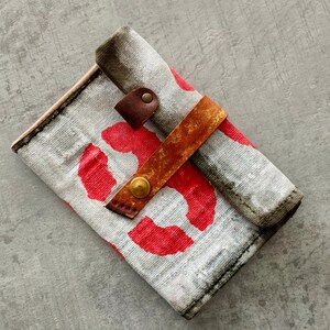 Könnte beinhalten: Eine handgefertigte Geldbörse mit einem grau-roten Muster. Die Geldbörse hat einen braunen Lederriemen mit einem Messingknopfverschluss. Die Kanten sind mit dunklem Faden genäht. Die Geldbörse ist geschlossen.