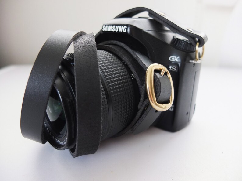 May include: A black camera with a black leather strap. The camera has a lens with a black lens cap. The camera has the word "SAMSUNG" printed on the top.