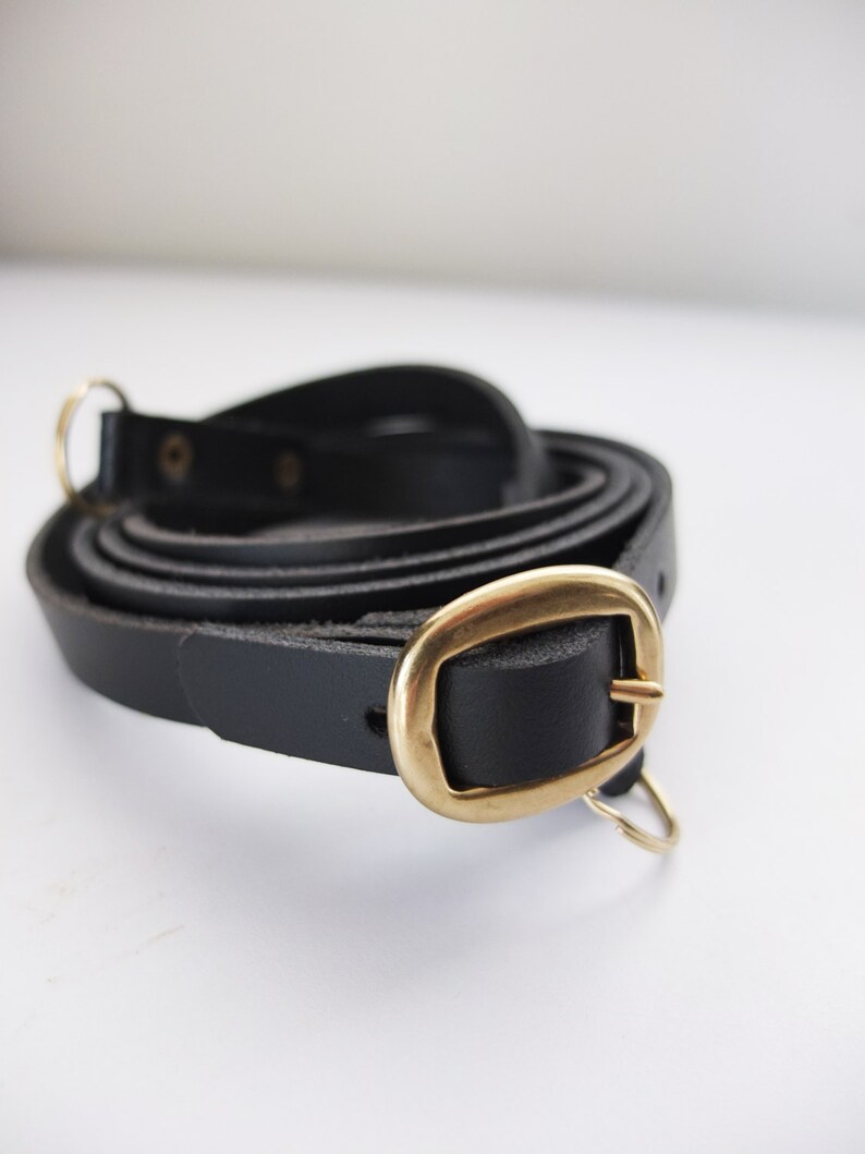 May include: A black leather camera strap with a gold metal buckle and two gold metal rings.