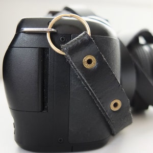 May include: A black camera with a leather strap and a gold ring attached to the strap. The camera has a black lens and a black body.