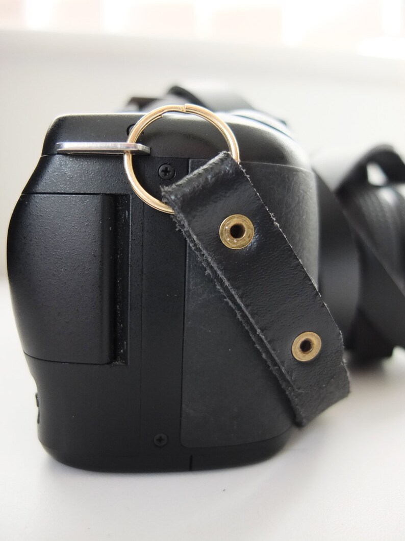 May include: A black camera with a leather strap and a gold ring attached to the strap. The camera has a black lens and a black body.