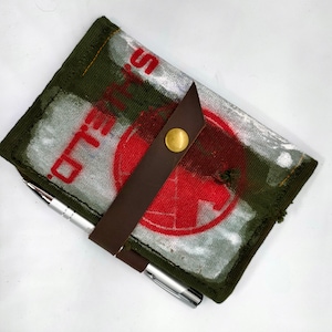 Può includere: Un taccuino di tela verde con un cerchio rosso e la parola "SOLD" in lettere bianche. Il taccuino ha un cinturino in pelle marrone con una chiusura a bottone in ottone. Una penna d'argento è appoggiata sul taccuino.