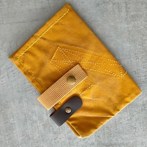 Può includere: Portafoglio in pelle giallo senape con un design cucito e una chiusura con cinturino intrecciato. Il portafoglio ha un cinturino in pelle marrone scuro con una chiusura a scatto in metallo. Le cuciture sono di una tonalità di giallo più chiara.