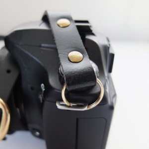 May include: A black camera with a black leather strap and gold metal accents. The strap has two gold metal rivets.