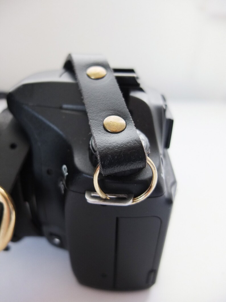 May include: A black camera with a black leather strap and gold metal accents. The strap has two gold metal rivets.