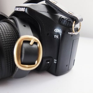 May include: A black camera with a leather strap and a gold buckle. The camera has a lens on the left side and a screen on the right side. The camera has the word "SAMSUNG" printed on the top.