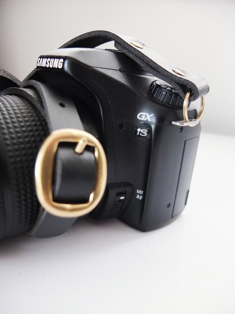 May include: A black camera with a leather strap and a gold buckle. The camera has a lens on the left side and a screen on the right side. The camera has the word "SAMSUNG" printed on the top.