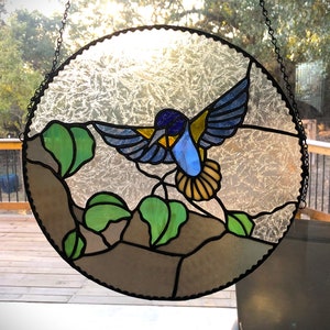 Puede incluir: Una ventana de vitral que representa un colibr&iacute; azul y amarillo con hojas marrones, verdes y blancas. El colibr&iacute; est&aacute; volando con las alas extendidas.