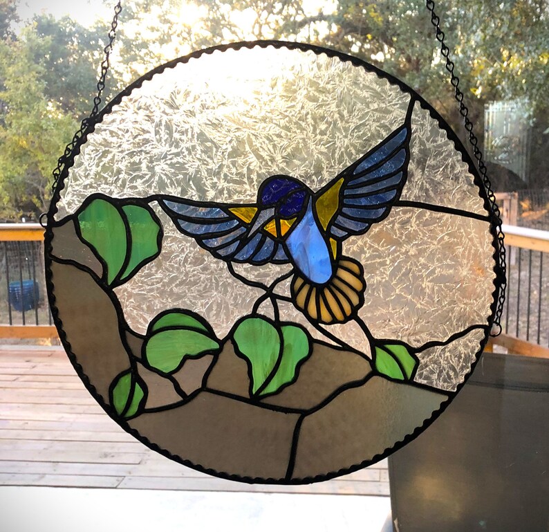 Puede incluir: Una ventana de vitral que representa un colibr&iacute; azul y amarillo con hojas marrones, verdes y blancas. El colibr&iacute; est&aacute; volando con las alas extendidas.