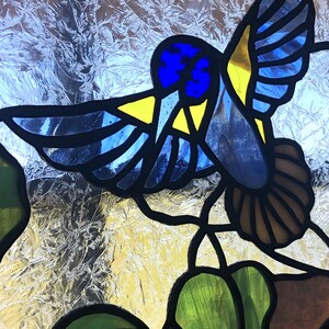Puede incluir: Un colibr&iacute; de vidrio emplomado con detalles azules, amarillos y marrones, posado en una hoja verde con un tallo marr&oacute;n.