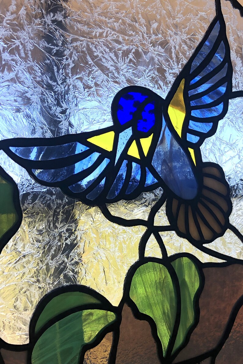 Puede incluir: Un colibr&iacute; de vidrio emplomado con detalles azules, amarillos y marrones, posado en una hoja verde con un tallo marr&oacute;n.