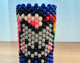 May include: A cylindrical beaded container in a vibrant array of colors. The design features blue, black, white, pink, gray, red, orange, yellow, green, and purple beads. The beaded pattern creates a face-like design. The container is standing on a wooden surface.