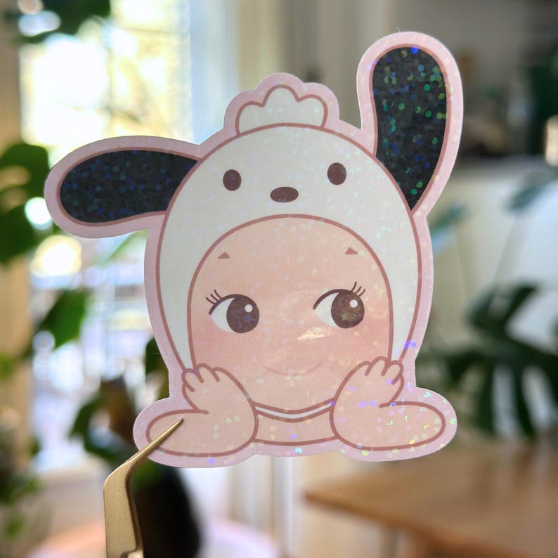 May include: A pink sticker featuring a baby wearing a white bunny hat with black glitter accents. The baby has brown eyes and is smiling. The sticker is held by a pair of tweezers.