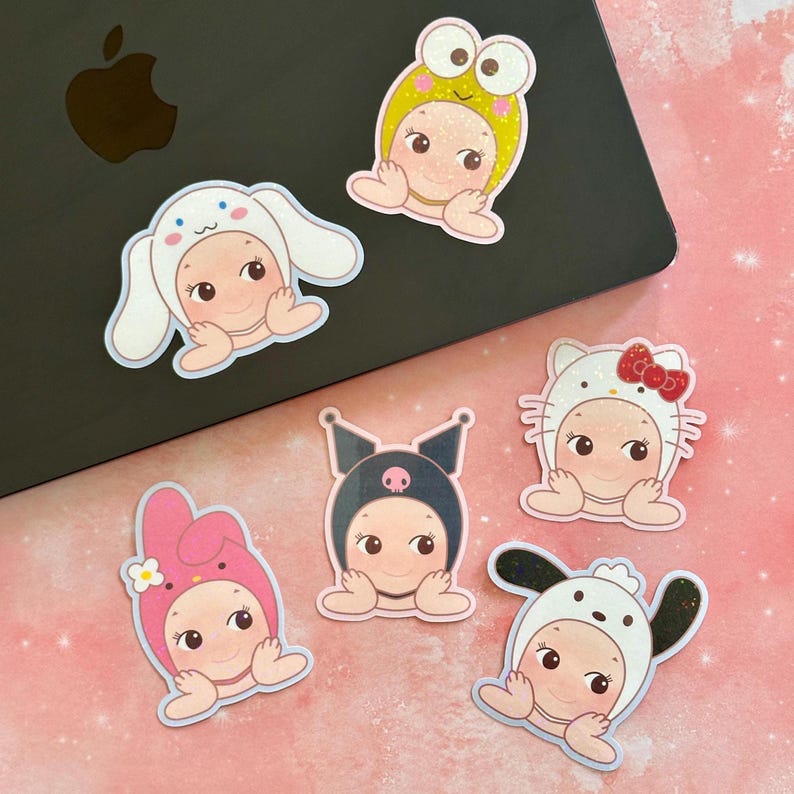 May include: A collection of six colorful stickers featuring adorable baby-faced characters in various animal and character-themed costumes. The stickers are on a black laptop and a pink surface.
