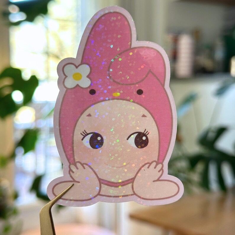 May include: A pink glitter sticker featuring a cartoon character in a bunny-like hood with a white flower accent. The character has large eyes and is holding its hands up near its face. The sticker is held by a pair of tweezers.