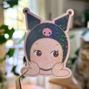 May include: A holographic sticker featuring a cartoon character with a pink face, large eyes, and a dark blue hood with a pink skull. The sticker has a glittery effect and is held by tweezers.
