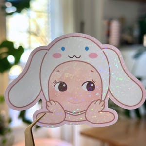 May include: A holographic sticker featuring a cartoon character in a bunny costume. The sticker has a pink and white color scheme with blue accents. The character has large eyes and is smiling. The sticker is held by tweezers.