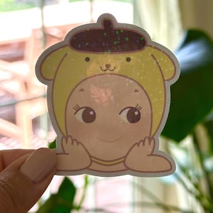 May include: A sticker featuring a cartoon character wearing a yellow dog hat with a brown beret. The character has large eyes and is smiling. The sticker has a holographic, glittery effect.