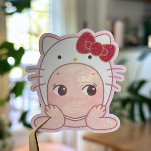 May include: A pink and white sticker featuring a baby in a Hello Kitty costume. The sticker has a glittery finish and a red bow. The baby has large brown eyes and is smiling. The sticker is held by a pair of tweezers.
