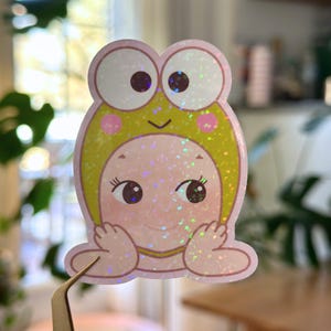 May include: A holographic sticker featuring a cartoon character in a frog hat. The sticker has a pink face, large eyes, and a yellow and green frog hat. The sticker is held by a pair of tweezers.