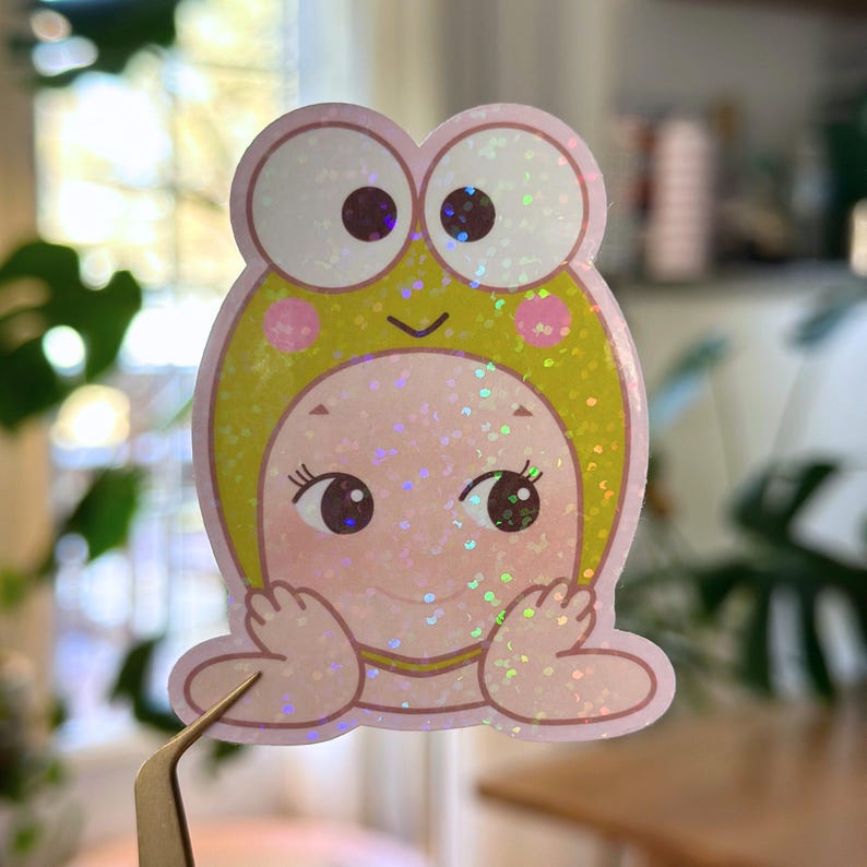 May include: A holographic sticker featuring a cartoon character in a frog hat. The sticker has a pink face, large eyes, and a yellow and green frog hat. The sticker is held by a pair of tweezers.