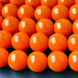 Puede incluir: Primer plano de una hebra de cuentas redondas de color naranja brillante. Las cuentas son lisas y brillantes.