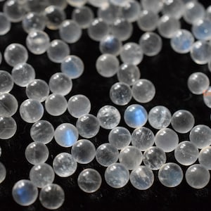 May include: A collection of round, white moonstone beads with a blue iridescent sheen. The beads are scattered on a black surface.