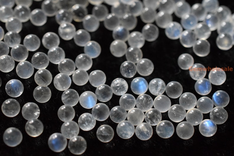 May include: A collection of round, white moonstone beads with a blue iridescent sheen. The beads are scattered on a black surface.