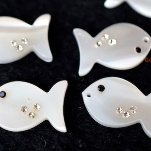 Może przedstawiać: Grupa pięciu białych muszlowych zawieszek w kształcie ryb z czarnymi i przezroczystymi cyrkoniami. Zawieszki mają około 5 cm długości i 2,5 cm szerokości.