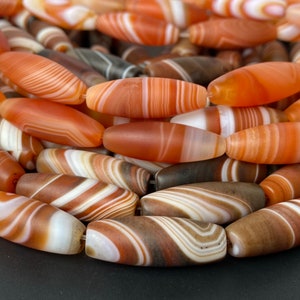 May include: A close-up of a collection of agate beads. The beads are oval shaped and have a variety of brown, white, and orange stripes.