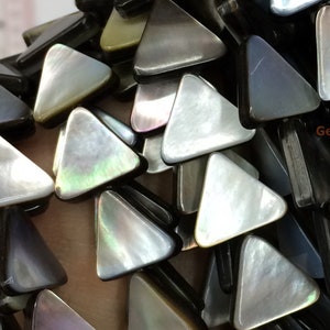 Puede incluir: Primer plano de un montón de cuentas de concha de forma triangular iridiscente. Las cuentas son una mezcla de negro, blanco y gris con un brillo nacarado. Las cuentas están dispuestas en un patrón aleatorio, con algunas superpuestas.