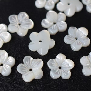 May include: A collection of white, mother-of-pearl flower-shaped beads with a hole in the center. The beads are carved with intricate details and have a glossy finish.