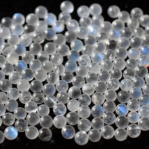 May include: A collection of round, white moonstone beads with a blue iridescent sheen. The beads are smooth and polished, and they are scattered on a black surface.