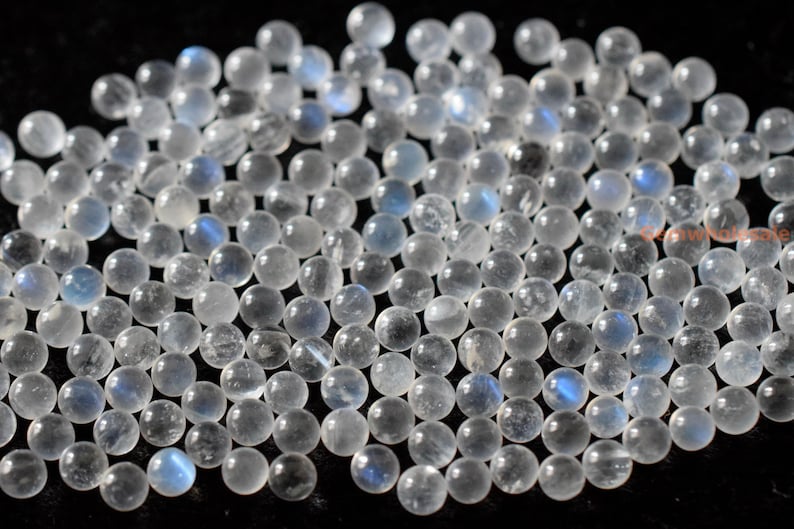 May include: A collection of round, white moonstone beads with a blue iridescent sheen. The beads are smooth and polished, and they are scattered on a black surface.