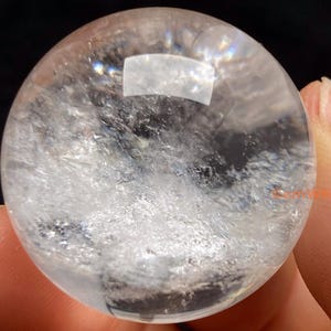 May include: A clear quartz crystal sphere, held between fingers, showcasing internal inclusions and a reflective surface. The sphere is transparent, allowing light to pass through, and is a popular item in the crystal and gemstone category.